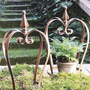 Può includere: Due portapiante a forma di cuore in metallo marrone arrugginito con volute decorative e un pinnacolo appuntito. Un supporto contiene una felce in vaso. I supporti sono su un letto di muschio verde, con uno sfondo sfocato di scale e fogliame.