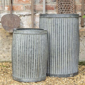 May include: Two weathered, grey metal planters with vertical ribbed detailing. The planters are cylindrical with a slightly wider midsection and a rim at the top and bottom. The background features a brick wall and gardening tools.