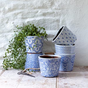 May include: A collection of blue and white ceramic plant pots with various patterns, including floral and geometric designs. One pot holds a green plant with trailing vines. The pots are stacked and arranged on a wooden surface, with a white brick wall in the background.