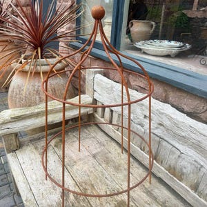 May include: A weathered, rusty brown metal plant support cage, featuring a domed top and a spherical finial. The cage has two circular bands and vertical supports. It is on a wooden bench.