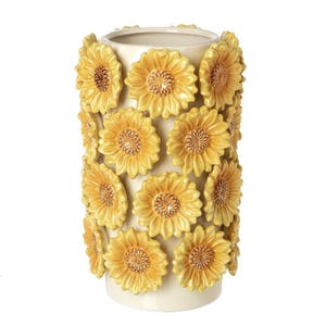 May include: A cream-colored ceramic vase adorned with dimensional yellow sunflowers. The vase is cylindrical, with the sunflowers arranged in rows around the exterior. The sunflowers have detailed centers and textured petals.