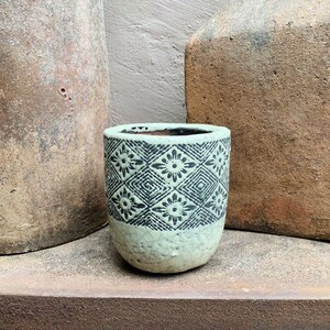 May include: A small, cylindrical ceramic planter featuring a light mint green base with a distinct crackle glaze finish. The upper portion is adorned with a dark charcoal grey geometric pattern consisting of repeating diamond shapes and floral motifs.