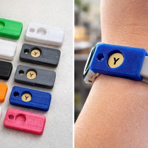 May include: A collection of colorful, rectangular watch band accessories. The accessories are in various colors, including green, white, black, gray, orange, blue, and pink. Each has a circular inset with a gold-colored disc bearing the letter "Y". One is shown attached to a watch.