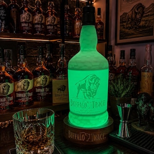 May include: A glowing green Buffalo Trace Kentucky Straight Bourbon Whiskey bottle lamp resting on a circular wooden base with the Buffalo Trace logo. The bottle features the brand's signature buffalo graphic and text, illuminated from within to emit a vibrant light.