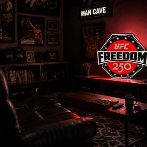 May include: A glowing, octagonal wall sign featuring the red, white, and black UFC Freedom 250 logo. The sign is illuminated against a dark wall, displaying the text UFC, FREEDOM, and 250 in a bold, stylized font within a circular border.