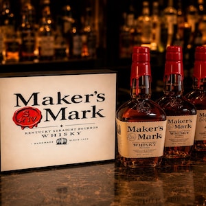 May include: A rectangular sign featuring the Maker's Mark logo and text alongside three glass bottles of Maker's Mark Kentucky Straight Bourbon Whisky. Each bottle has a distinctive red wax seal on the neck and a cream-colored paper label.