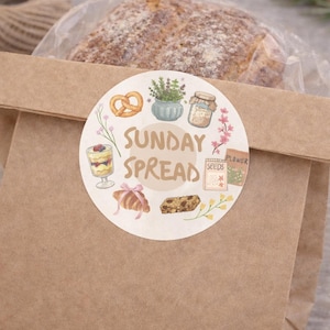 May include: A brown paper bag with a loaf of bread inside a clear wrapper. The bag has a circular sticker with the words "SUNDAY SPREAD" and illustrations of food items, including a pretzel and a croissant.