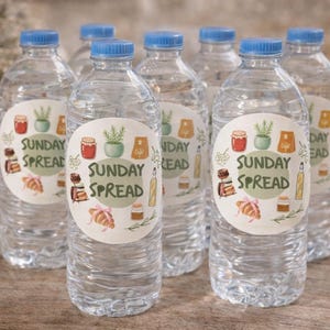Puede incluir: Varias botellas de agua transparentes con tapas azules. Cada botella tiene una etiqueta circular con las palabras "SUNDAY SPREAD" e ilustraciones de alimentos. Las botellas están dispuestas sobre una superficie de madera.