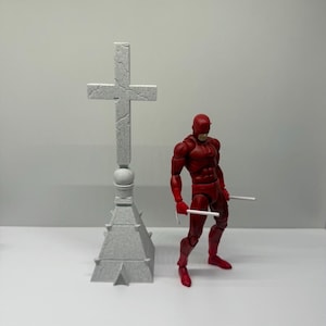 May include: A red action figure with a mask and two white batons standing next to a tall, gray, stone-textured cross monument with a pyramid-shaped base. The cross features visible cracks and a weathered appearance on its surface.