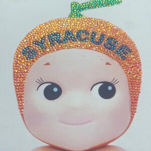 May include: A whimsical illustration of a baby-like figure with an orange headpiece adorned with sparkling rhinestones. The word "SYRACUSE" is spelled out in blue rhinestones across the headpiece. The figure has large eyes and a gentle smile.