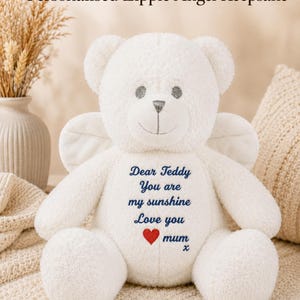 Könnte beinhalten: Ein weißer Plüsch-Teddybär mit Engelsflügeln auf dem Rücken. Der Bär hat gestickte graue Augen und eine Nase sowie blauen Text auf der Brust: Dear Teddy You are my sunshine Love you, gefolgt von einem roten Herz, dem Wort mum und einem x.