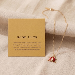 以下が含まれることがあります： 「GOOD LUCK」と書かれたカードと一緒に展示されている、赤と黒のてんとう虫のペンダントが付いたゴールドカラーのネックレス。 てんとう虫のペンダントは、黒い斑点と金のアクセントが付いています。 カードには幸運と願い事に関するテキストが含まれています。