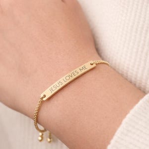 Puede incluir: Pulsera dorada con una barra rectangular grabada con las palabras "JESUS LOVES ME". La pulsera está en una delicada cadena con cuentas ajustables. La pulsera se lleva en la muñeca.