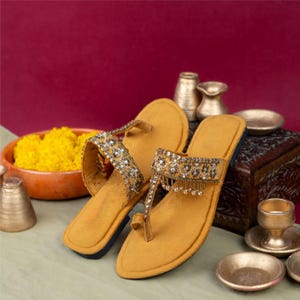 May include: Mustard yellow sandals with intricate beading and dangling embellishments. The sandals feature a toe loop design and are adorned with sparkling rhinestones. The footwear is displayed on a light green surface with a maroon background, suggesting a focus on fashion and accessories.