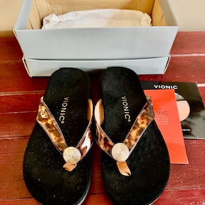 May include: Pair of black Vionic flip-flops with brown and tan patterned straps and a gold-colored circular embellishment. The sandals are displayed with their original box and a product information card.