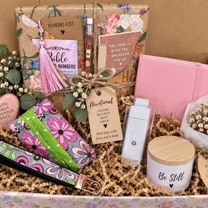 May include: A gift box containing a pink notebook, a white candle with a wooden lid labeled Be Still, a white book light, a floral-wrapped item, a pink tassel, a heart-shaped stone with text, a floral wristlet, and a tag reading Devotional Inspire Reflect Pray Grow.