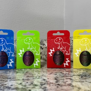 May include: Four colorful boxes, blue, green, red, and yellow, each with a dinosaur outline and a glittery egg-shaped object. The boxes have a zig-zag design and a hole at the top.