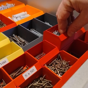 Può includere: Primo piano di un organizzatore di stoccaggio multicolore pieno di vari articoli di ferramenta. L'organizzatore ha scomparti arancioni, gialli e grigi, ciascuno con viti, bulloni e tasselli. Una mano raggiunge uno degli scomparti arancioni.
