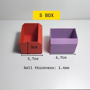 May include: Two small, open-top storage boxes. One is red and measures 5 cm high and 5.7 cm wide. The other is purple and is 6.7 cm wide. The text "S BOX" is in a yellow rectangle above the boxes.
