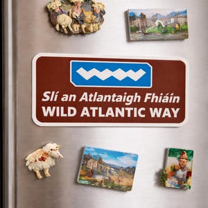 May include: A rectangular magnet featuring the brown and white Wild Atlantic Way road sign with the text Slí an Atlantaigh Fhiáin, displayed alongside other decorative magnets including sheep figures and landscape scenes on a metal surface.