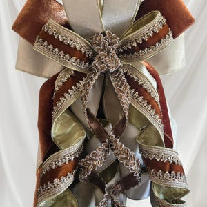May include: A decorative bow featuring layered ribbons in rust-colored velvet and champagne-colored satin. The edges are adorned with intricate metallic gold and silver beaded trim, with additional braided metallic ribbon accents cascading down.