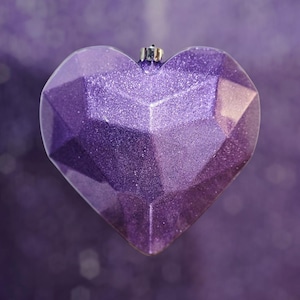 May include: A faceted, heart-shaped ornament in shades of purple, with a glittery finish. The ornament has a silver metal cap for hanging. The background is a blurred, matching purple.