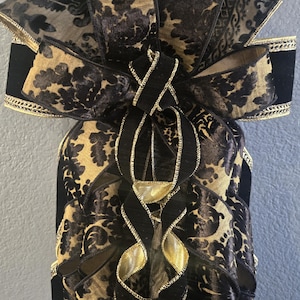 May include: A decorative bow crafted from black velvet and gold-toned fabric featuring a dark damask pattern. The ribbon edges are finished with metallic gold trim, and the design includes multiple cascading loops and long, flowing tails.