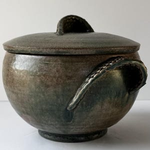 以下が含まれることがあります： 濃い緑と茶色のまだら模様の釉薬が施された、蓋付きの丸い陶器の壺。壺の側面には湾曲した取っ手があり、蓋の上部には質感のあるアーチ型の取っ手が付いています。表面は滑らかで、土のような風合いのある仕上がりになっています。