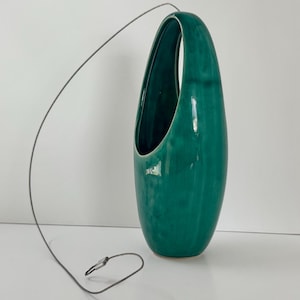Op de afbeelding: Een traanvormige, blauwgroene keramische wandplantenbak met een glanzende afwerking en een grote ovale opening. Een dunne, flexibele grijze metalen draad is aan de bovenkant bevestigd en loopt in een boog naar een kleine metalen sluiting onderaan.