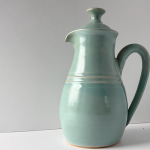 Può includere: Una brocca in ceramica color azzurro con finitura lucida, caratterizzata da un corpo arrotondato, un manico curvo, un beccuccio e un coperchio abbinato con pomello. Il design presenta sottili scanalature orizzontali sul collo del vaso.