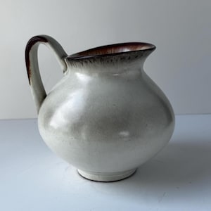 May include: A ceramic pitcher with a rounded body and a curved handle. The exterior features a smooth, off-white glaze with subtle grey shading. The rim and the top of the handle are accented with a dark brown, drip-style glaze finish.