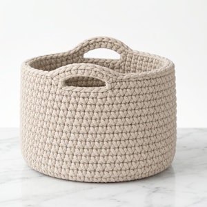 May include: A beige, round crochet basket with two integrated handles. The basket is made of thick yarn and has a textured appearance. The neutral color and simple design make it suitable for various storage needs.
