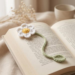 May include: A handmade crochet daisy bookmark with white petals and a yellow center, resting on the pages of an open book. The bookmark has a green stem and leaf, perfect for book lovers.