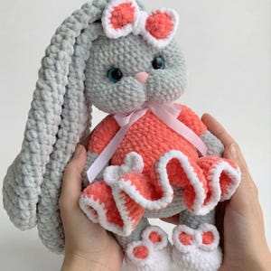 May include: A handmade gray plush bunny with long ears, wearing a coral dress with white trim and a matching bow. The bunny has blue eyes and a pink nose, and is held in someone's hands.