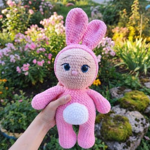 May include: A handmade pink crochet bunny doll with large blue eyes and a white fluffy tail. The bunny has long ears and is wearing a hood. The doll is held in front of a background of pink flowers and greenery.