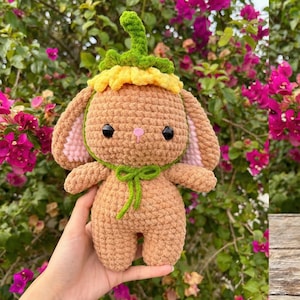 May include: A handmade crochet bunny doll with a flower hat. The bunny is tan with pink inner ears, black eyes, and a green bow. The hat is yellow and green. The doll is held in front of a background of pink flowers.