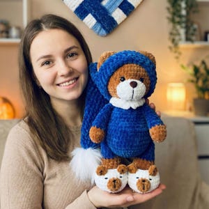 Pode incluir: Um urso de pelúcia castanho feito à mão usando um traje azul, chapéu e chinelos a condizer. O urso tem uma gola branca e um pompom branco no chapéu. Os chinelos do urso têm um design de rosto de urso.