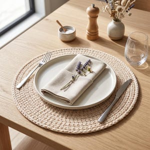 May include: A beige crocheted placemat with a white plate, linen napkin, and silverware set on a wooden table. The napkin is adorned with lavender and small yellow flowers. A glass, salt cellar, and pepper grinder are also present.