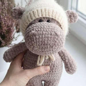 May include: A handmade, plush hippo toy in shades of gray and cream. The hippo wears a cream-colored knitted hat and a small cream-colored scarf. The toy is soft and cuddly, perfect for a child's room or nursery.