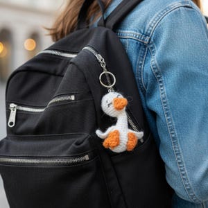 May include: A white and orange crochet goose keychain attached to a black backpack. The goose has closed eyes and outstretched wings. The backpack has silver zippers and is worn with a denim jacket.