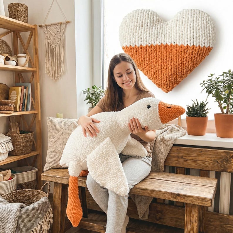 May include: A large, plush white goose with orange beak and feet is held by a person. A large, heart-shaped wall hanging in white and orange is visible. The scene is set in a cozy, well-lit room with wooden furniture and plants.