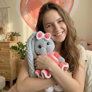 Può includere: Un coniglietto di peluche fatto a mano in grigio tenue, corallo e bianco. Il coniglietto ha lunghe orecchie flosce, un fiocco color corallo e un vestito color corallo con finiture bianche. Il coniglietto è tenuto da una persona.