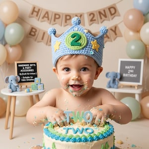 May include: A baby wearing a light blue crochet crown with yellow stars and a green number 2. The baby is covered in cake frosting and holding a cake with the word "TWO" on top. A sign reads "HANDMADE GIFTS & PARTY ACCESSORIES".
