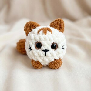 May include: A handmade crochet cat toy. The toy features a white body with brown accents on the ears, paws, and a stripe on the head. It has large, dark eyes with a glittery gold outline and a small black nose and whiskers. The toy is soft and plush.