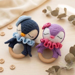 Op de afbeelding: Twee handgemaakte gehaakte baby rammelaars in de vorm van een pinguïn. Eén is marineblauw met een lichtblauwe hoed en ruches, de andere is paars met een roze strik en ruches. Beide hebben houten ringhandvatten en gesloten ogen.