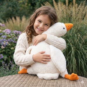 May include: A plush white goose toy with an orange beak and feet is being hugged. The goose has a soft, textured body. The toy is on a wicker table, with a background of greenery and flowers.