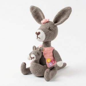 May include: A crocheted kangaroo and joey in grey, white, and pink. The larger kangaroo wears a pink vest with colourful flower appliqués. The joey sits in the mother's pouch. The kangaroo has a small pink bow on its head.