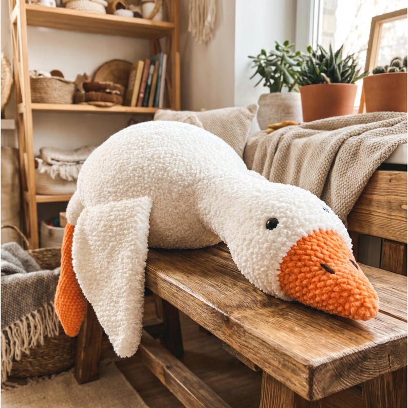 May include: A large, plush white goose with orange beak and feet, resting on a wooden bench. The soft, textured toy has black eyes and is designed to resemble a real goose. The plush toy is a decorative item.