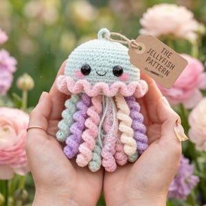 Pode incluir: Um brinquedo de água-viva de crochê em cores pastel. A água-viva tem uma cabeça azul clara, babados rosa e branco e tentáculos em espiral em tons de rosa, roxo e verde. Uma etiqueta diz "THE JELLYFISH PATTERN".