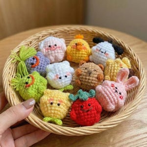 Puede incluir: Una colección de adornos coloridos con forma de animales tejidos a ganchillo, incluyendo un cerdo, un pollito, un perro, un oso y un conejo, dispuestos en una cesta de mimbre. Los adornos son de varios colores, como rosa, amarillo, blanco y marrón, con detalles bordados.